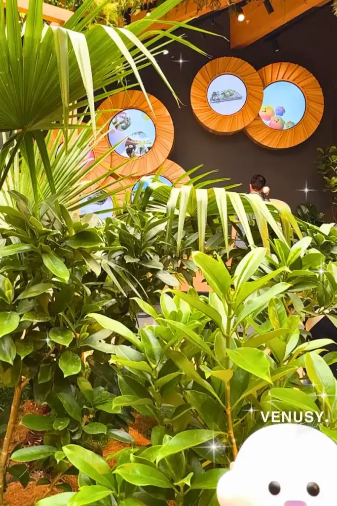 大阪・関西万博ドイツ館のグリーンエリアで、植物の間から顔をのぞかせるサーキュラーちゃんの様子。背景には循環型素材の展示も見える。