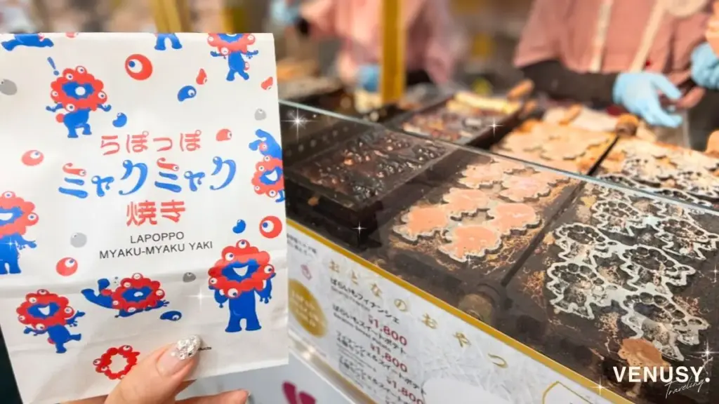 ミャクミャク焼き紙袋と実演シーン：ヴィナぽ♡が手にした「らぽっぽミャクミャク焼き」の公式袋と、奥で焼かれているミャクミャク焼きのライブ感が同時に写った1枚。
