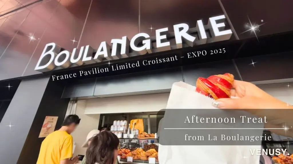 大阪万博フランス館の「La Boulangerie」で購入した限定クロワッサンを手に持ち、店舗外観を背景に撮影した写真